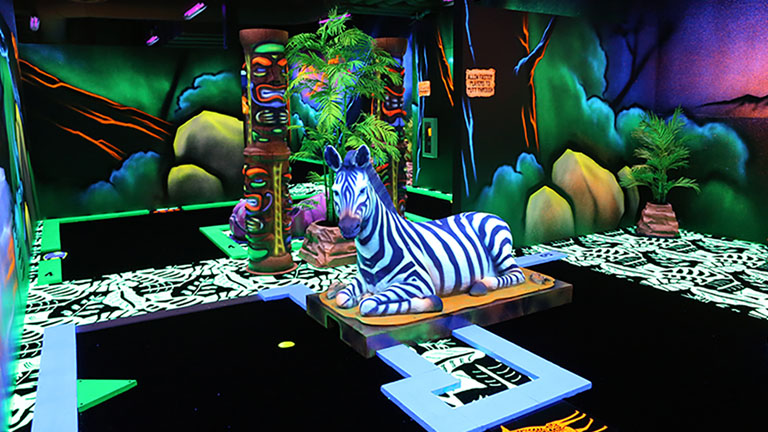An overview of our blacklight mini golf course with a sitting zebra sculpture and African landscape..